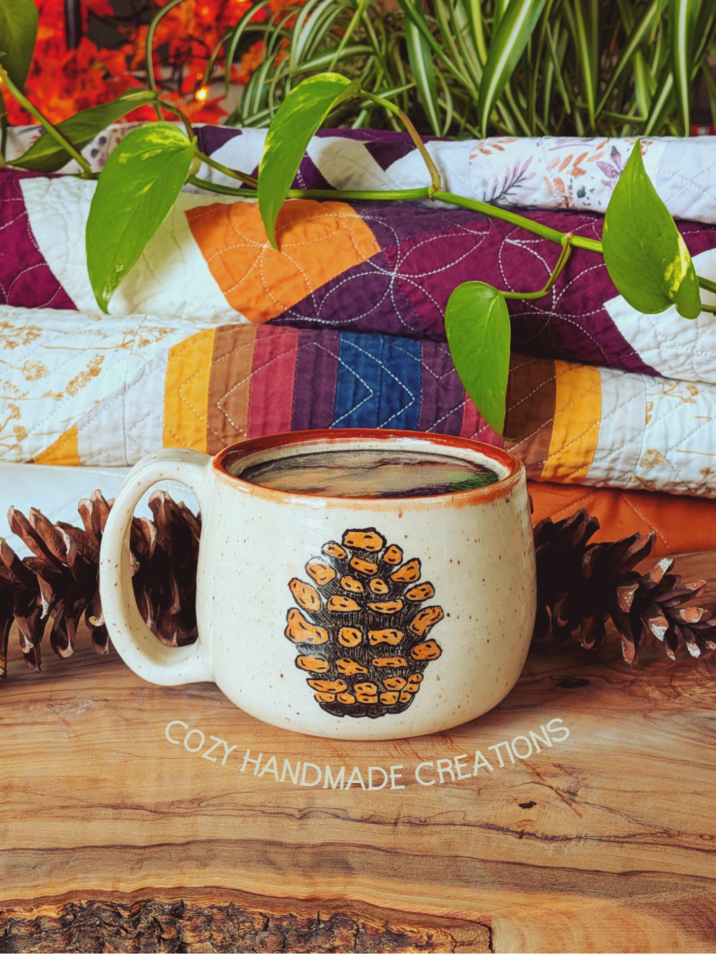 White mug with a pine cone design on a wooden surface, surrounded by plants and fabric.